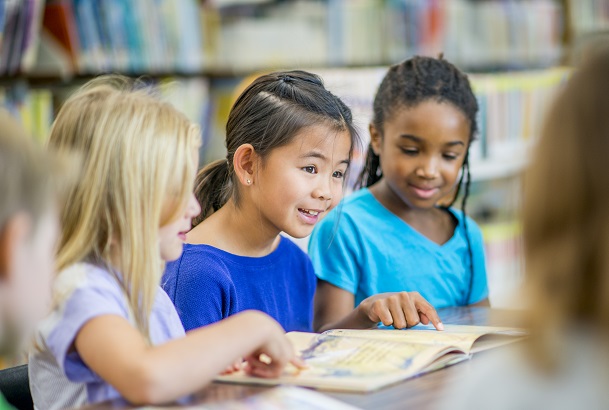 A group of elementary school girls of different ethnicity are indoors in their school library. They are sitting together at a table and reading together.
