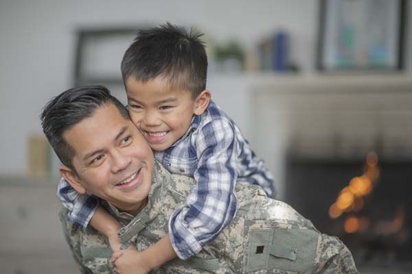 A military dad and his son are hugging in their living room. The son is smiling happily at the camera.